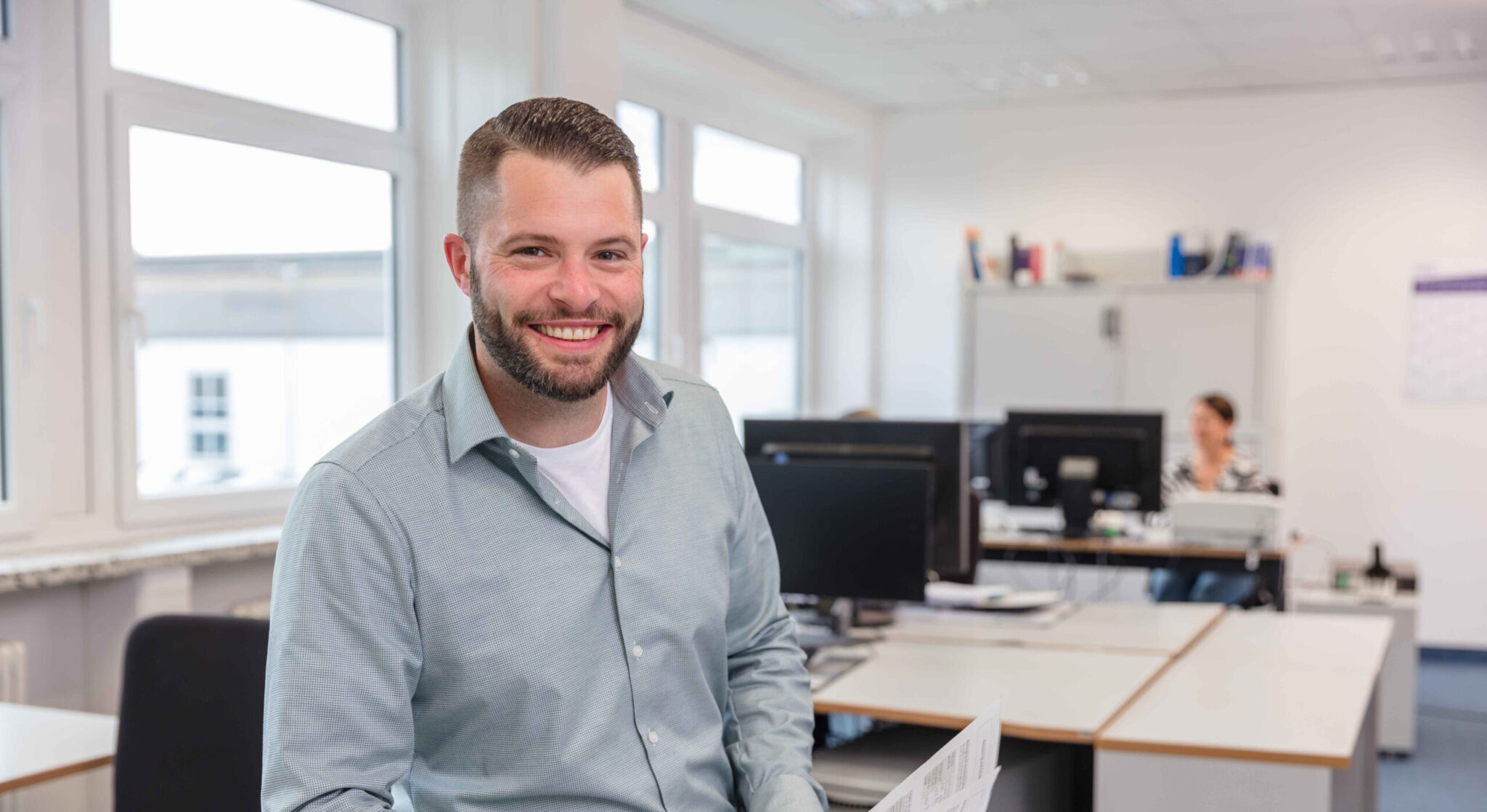REINTJES Mitarbeiter im Büro, während eine andere Mitarbeiterin im Hintergrund am Schreibtisch zu erkennen ist.